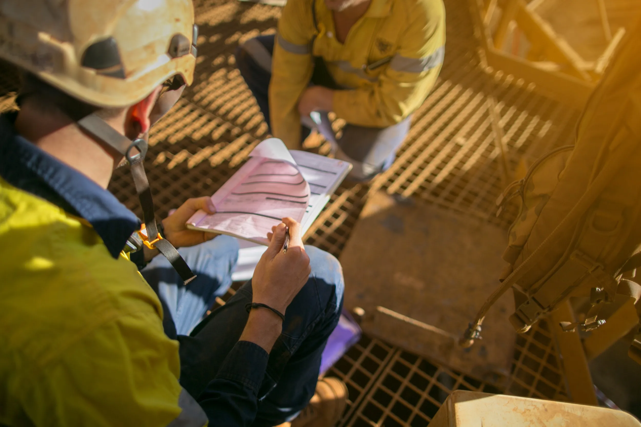 Two workers in helmets and high-visibility clothing crouched on an elevated steel grating, reviewing notes